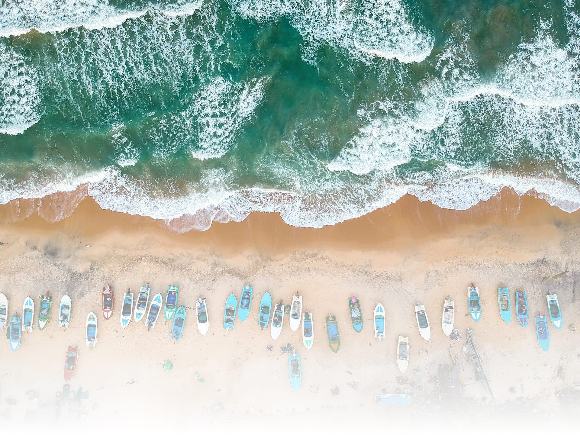 Fishing boats drawn up in a line on the sand where the surf breaks, seen from directly above